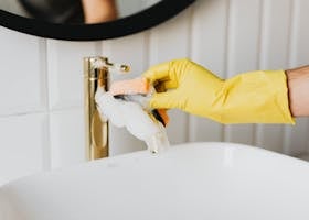 Closeup of a hand in a yellow glove cleaning a bathroom faucet with a sponge and soap.